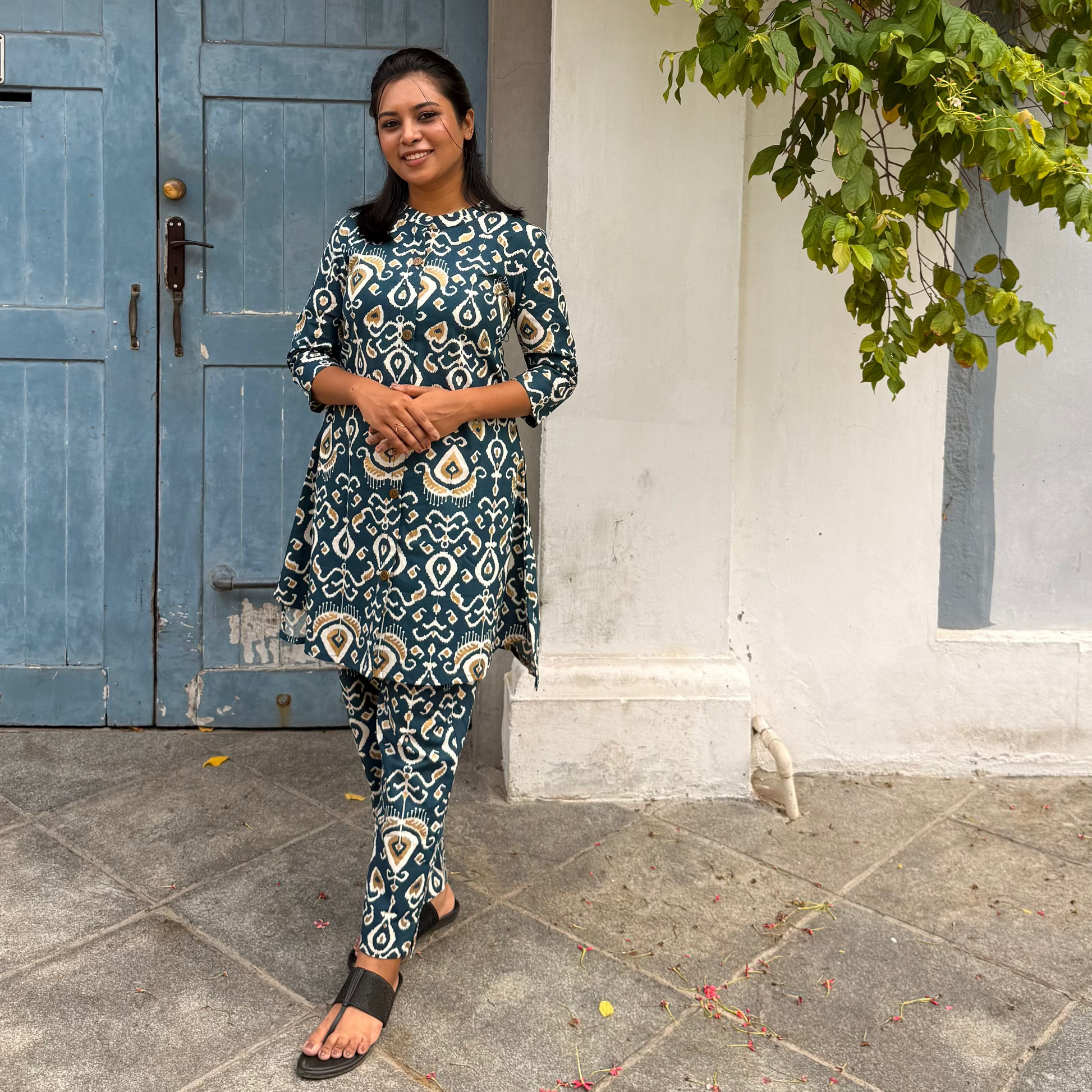 Woman in a patterned outfit standing in front of a blue door with greenery above.
