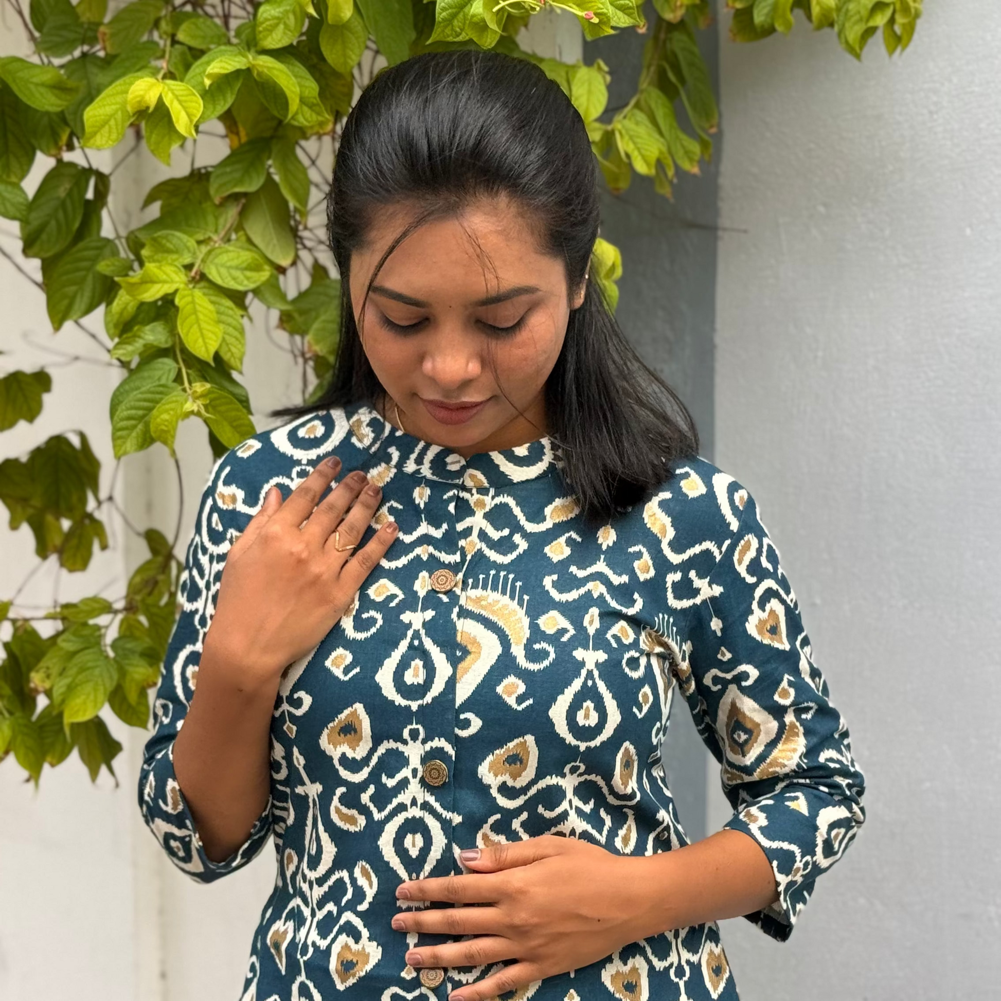 Woman wearing a blue patterned dress standing against a white wall with green leaves.